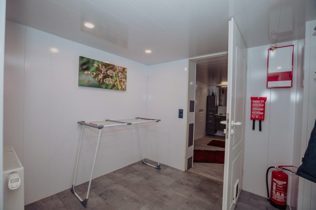 Flur im Nebeneingangsbereich mit Garderobe und Schuhablage. Eine Tür führt direkt hinaus in den geschützten Innenhof. Der Boden ist hell.