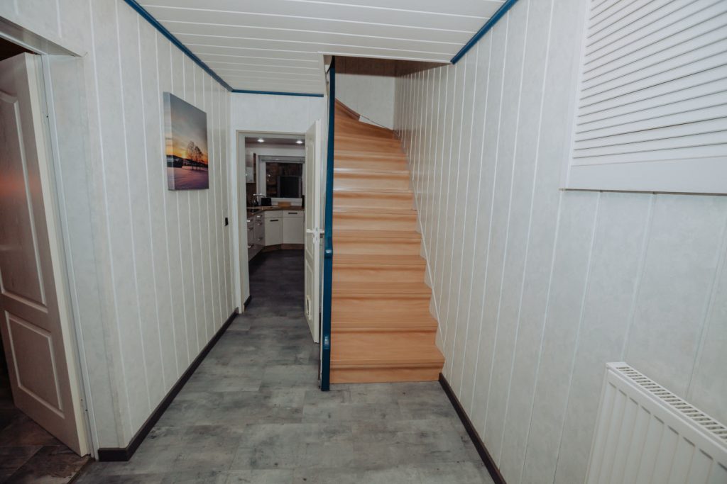 Heller Flur im Erdgeschoss mit grauer Bodenoptik und weißen Wandpaneelen. Eine Holztreppe führt nach oben. Durch die offene Tür im Hintergrund blickt man direkt in die moderne Küche.