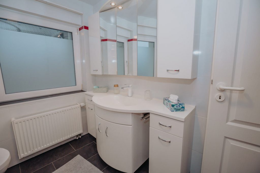 Helles Badezimmer mit weißem Waschtisch, großem dreitürigen Spiegelschrank und Fenster mit Milchglas-Sichtschutz. Dunkle Bodenfliesen bilden einen Kontrast zu den weißen Möbeln.