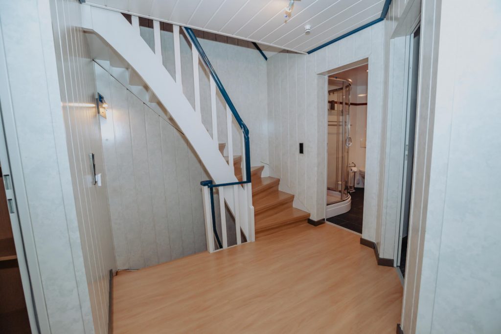 Helle Diele in der ersten Etage mit Laminatboden. Eine weiße Holztreppe mit blauem Handlauf führt nach oben. Rechts sieht man durch die offene Tür in das Badezimmer mit Duschkabine.