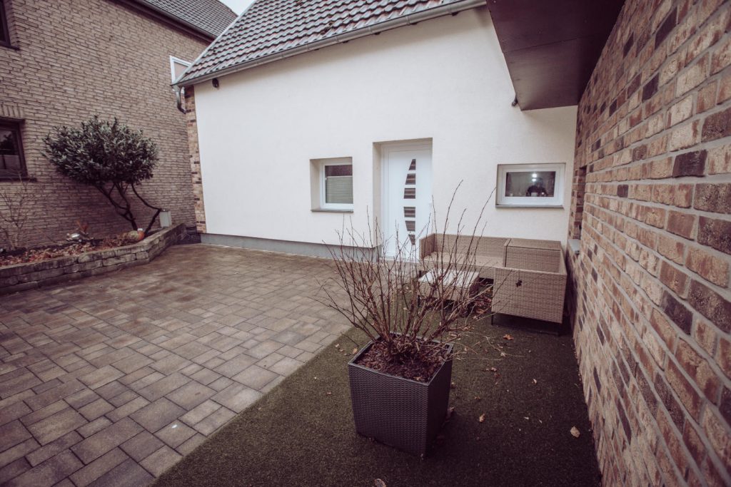 Gepflasterter Innenhof mit weißer Nebeneingangstür. Rechts an der Klinkerwand steht eine Rattan-Sitzgruppe (Lounge-Möbel) mit Tisch. Im Vordergrund ein großer grauer Pflanzkübel