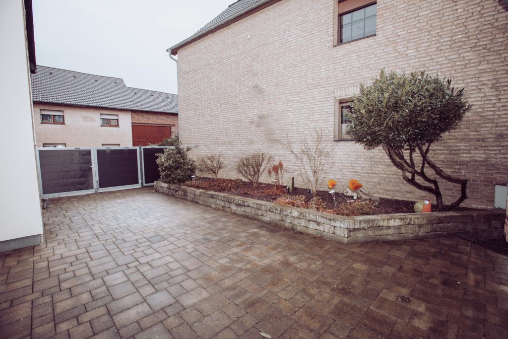 Gepflasterter Innenhof mit einem breiten, undurchsichtigen Tor zur Straße hin. Rechts ein gemauertes Hochbeet mit Pflanzen und einem kleinen Baum.
