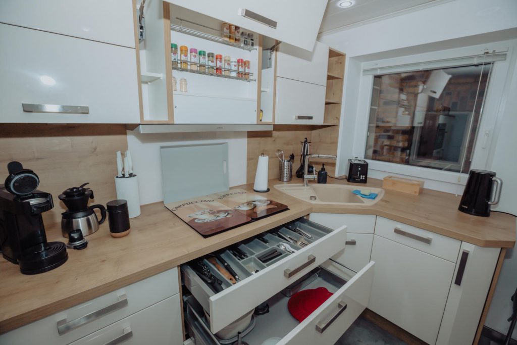 Geöffnete Küchenschubladen mit vollem Besteckkasten und Kochutensilien. Auf der Arbeitsfläche darüber stehen Wasserkocher, Toaster und Kaffeemaschinen vor einem Fenster.
