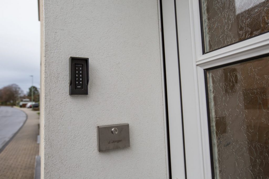 Weißer Hauseingang mit einem schwarzen Nuki Keypad an der Wand für den schlüssellosen Zugang. Darunter eine Klingel aus Edelstahl. Im Hintergrund ist eine ruhige Dorfstraße zu sehen.