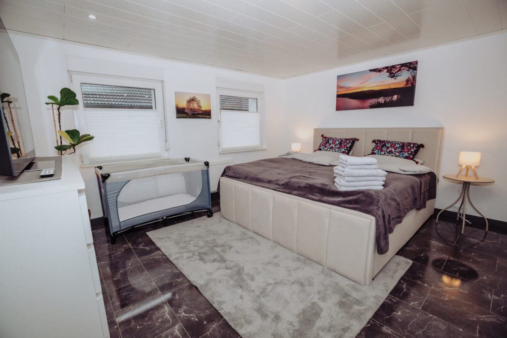 Helles Schlafzimmer mit großem Doppelbett und einem aufgebauten Baby-Reisebett. Auf dem Bett liegen frische Handtücher bereit. An der Wand hängen Landschaftsbilder.