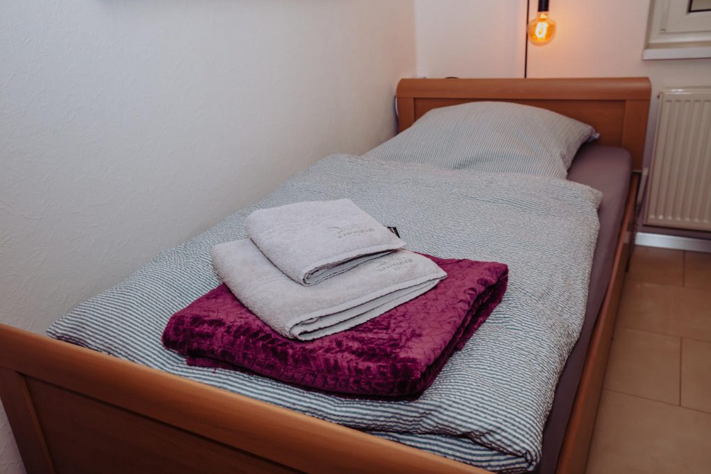Einzelbett mit Holzrahmen und grau-weiß gestreifter Bettwäsche. Darauf liegen eine gefaltete violette Kuscheldecke und zwei frische graue Handtücher. Im Hintergrund leuchtet eine warme Hängelampe.