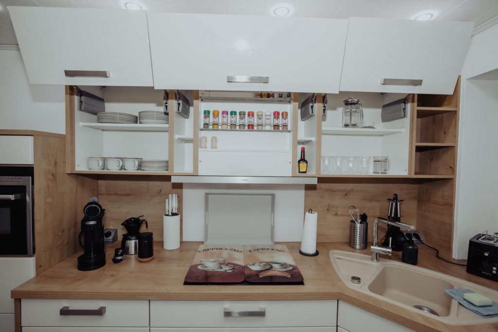 Geöffnete Küchen-Oberschränke gefüllt mit weißem Geschirr, Gläsern und einem vollen Gewürzregal. Darunter eine Arbeitsplatte mit Eckspüle, Kaffeemaschine und Messerblock.