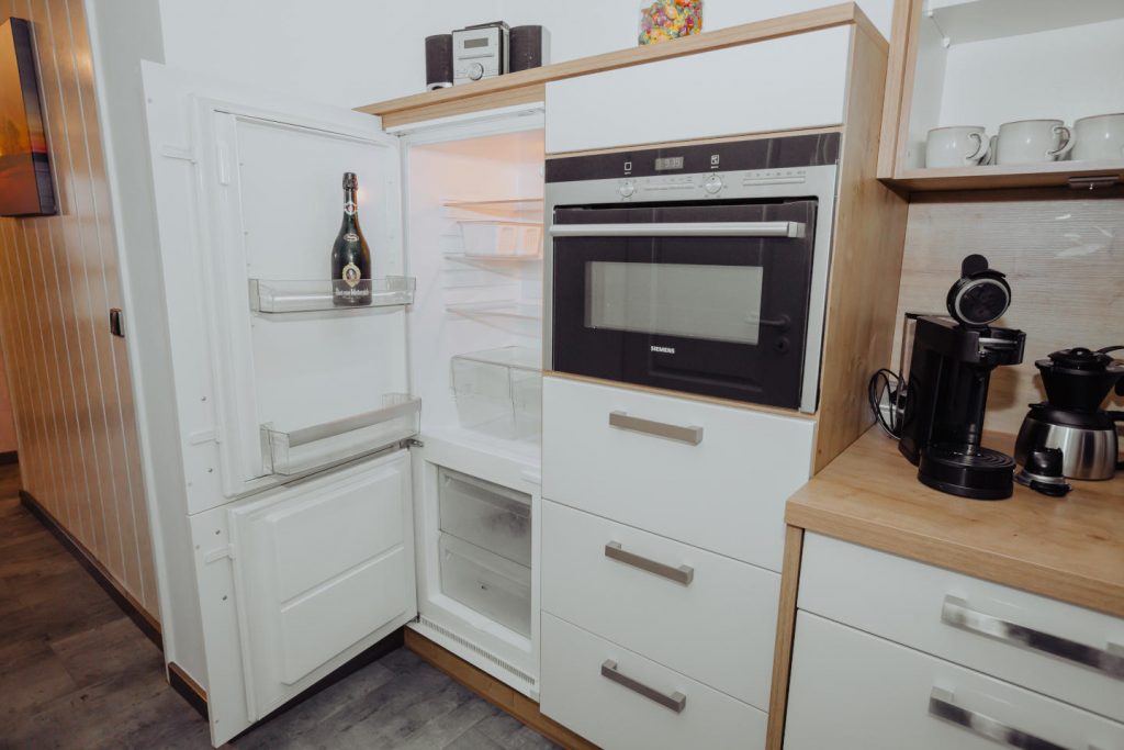 Offener, integrierter Kühlschrank mit einer Flasche Sekt. Daneben ein Einbau-Backofen der Marke Siemens, eine Kapselkaffeemaschine und eine Filterkaffeemaschine auf einer Holz-Arbeitsplatte in einer modernen Küche.