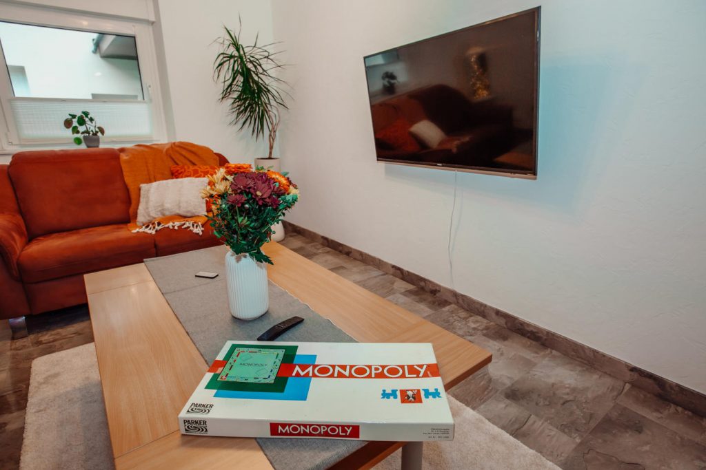 Gemütliches Wohnzimmer im Ferienhaus mit Sofa, Smart-TV an der Wand und Monopoly-Spiel auf dem Couchtisch